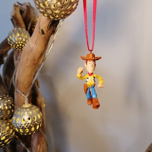 May include: A plastic toy ornament of Woody from Toy Story, wearing his signature cowboy hat and vest. He is holding his arm out in a gesture of excitement. The ornament is hanging from a red ribbon.
