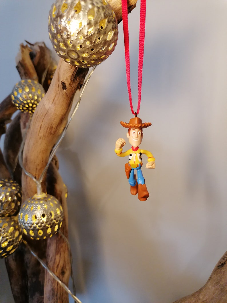 May include: A plastic toy ornament of Woody from Toy Story, wearing his signature cowboy hat and vest. He is holding his arm out in a gesture of excitement. The ornament is hanging from a red ribbon.