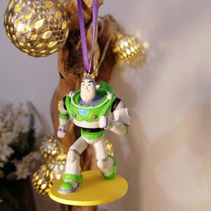 K&ouml;nnte beinhalten: Ein gr&uuml;ner und wei&szlig;er Plastik-Anh&auml;nger mit Buzz Lightyear aus der Toy Story-Reihe. Der Anh&auml;nger h&auml;ngt an einem lila Band und hat eine gelbe Basis.