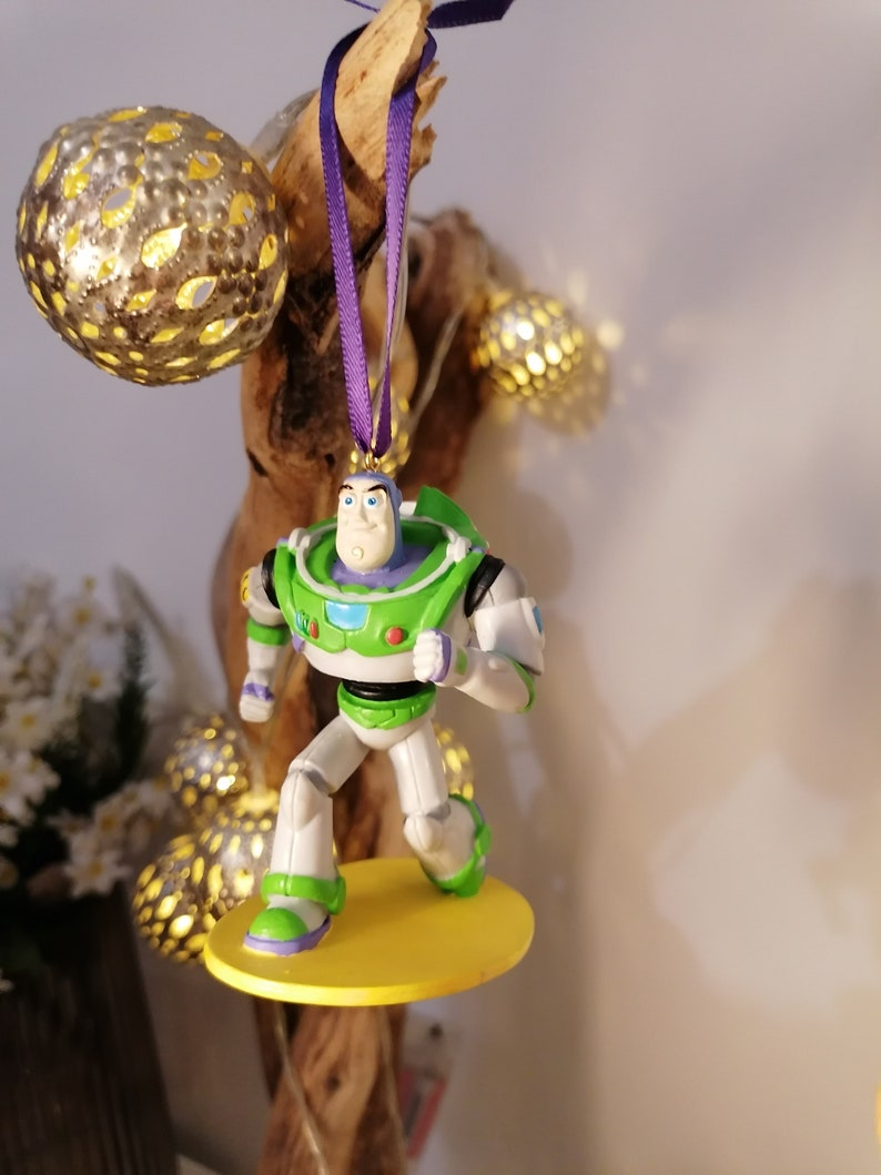 K&ouml;nnte beinhalten: Ein gr&uuml;ner und wei&szlig;er Plastik-Anh&auml;nger mit Buzz Lightyear aus der Toy Story-Reihe. Der Anh&auml;nger h&auml;ngt an einem lila Band und hat eine gelbe Basis.