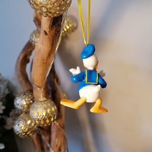 Op de afbeelding: Een blauwe en gele Donald Duck kerstversiering die aan een tak hangt met een geel lint. De versiering is gemaakt van plastic en heeft een gedetailleerd ontwerp.