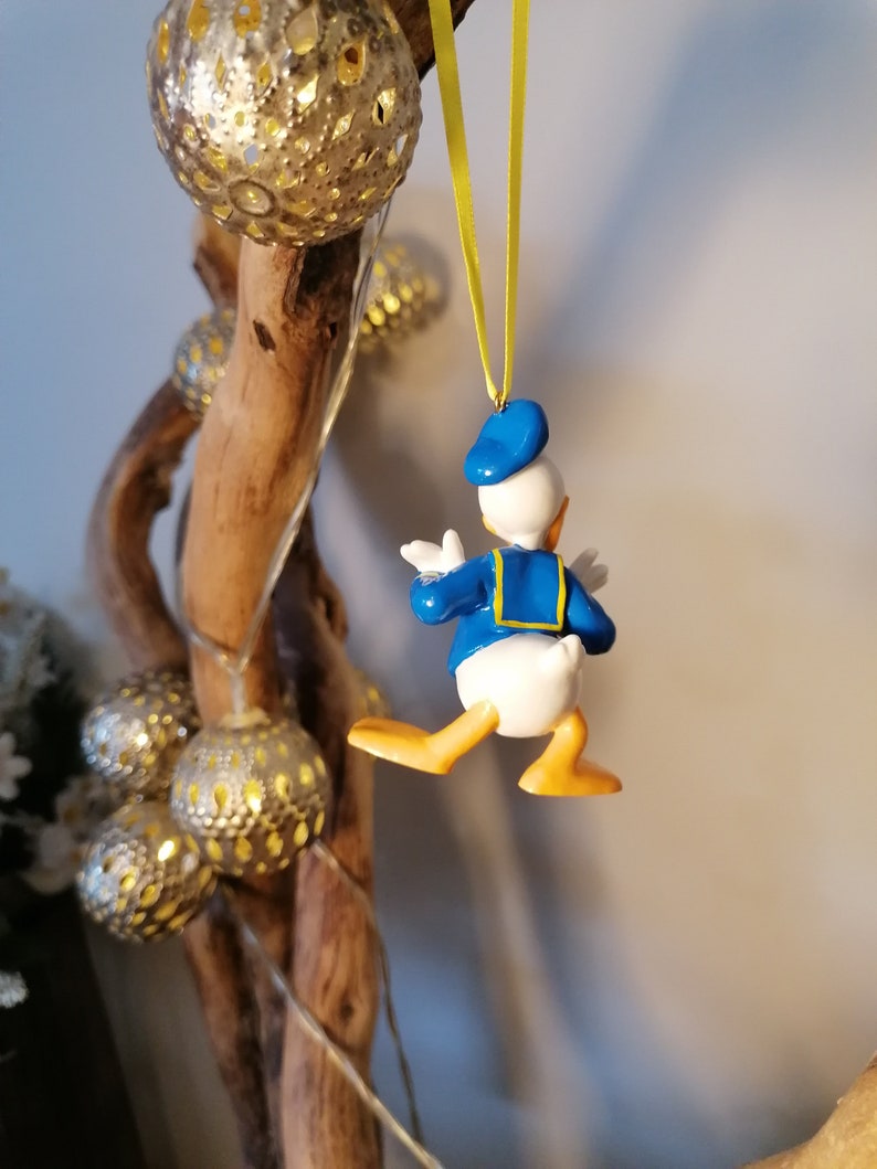 Op de afbeelding: Een blauwe en gele Donald Duck kerstversiering die aan een tak hangt met een geel lint. De versiering is gemaakt van plastic en heeft een gedetailleerd ontwerp.