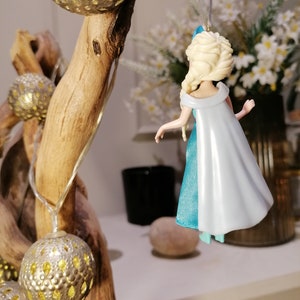 May include: A blue and white plastic figurine of a female character with blonde hair and a blue and white dress. The figurine is hanging from a string.