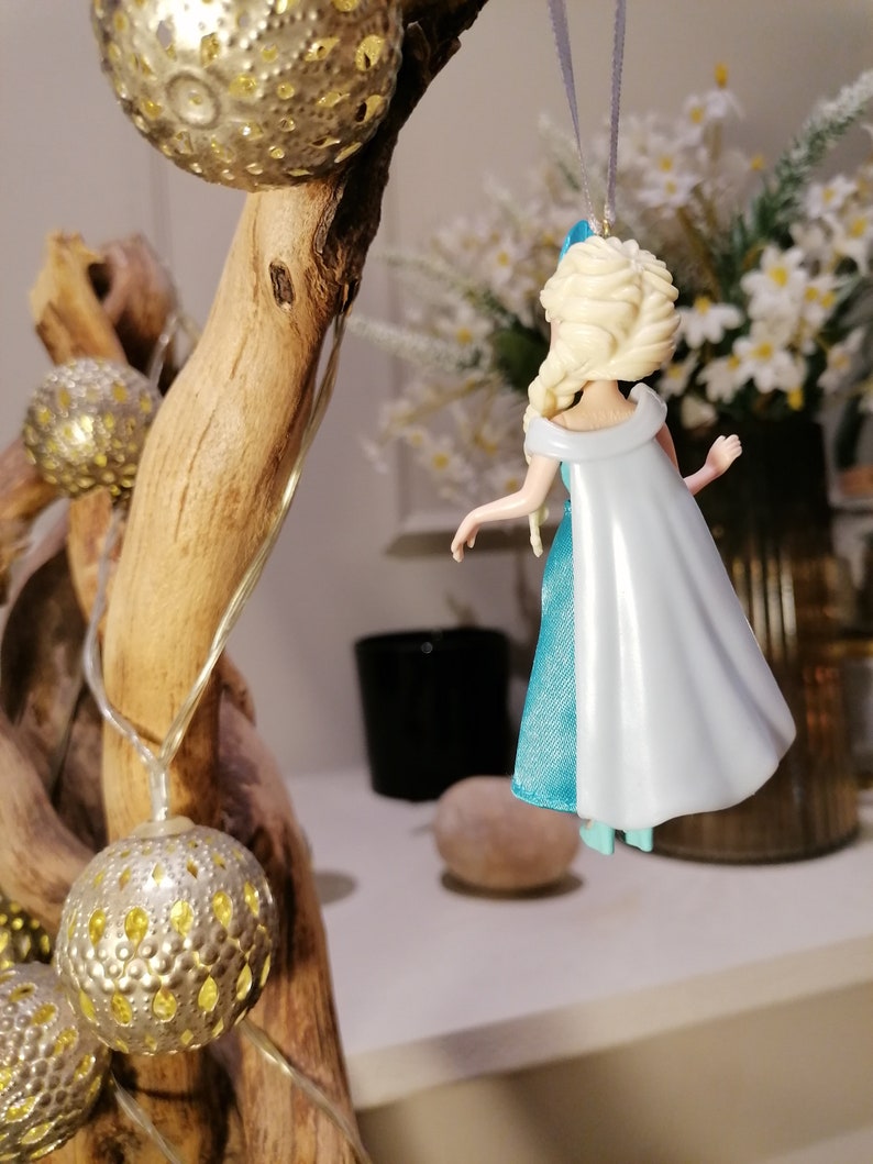 May include: A blue and white plastic figurine of a female character with blonde hair and a blue and white dress. The figurine is hanging from a string.