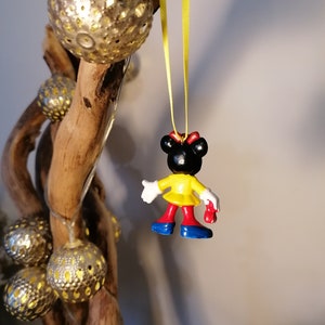K&ouml;nnte beinhalten: Eine kleine Plastikfigur eines Comic-Charakters, die ein gelbes Kleid, rote Schuhe und eine rote Schleife tr&auml;gt. Die Figur h&auml;ngt an einem gelben Band.