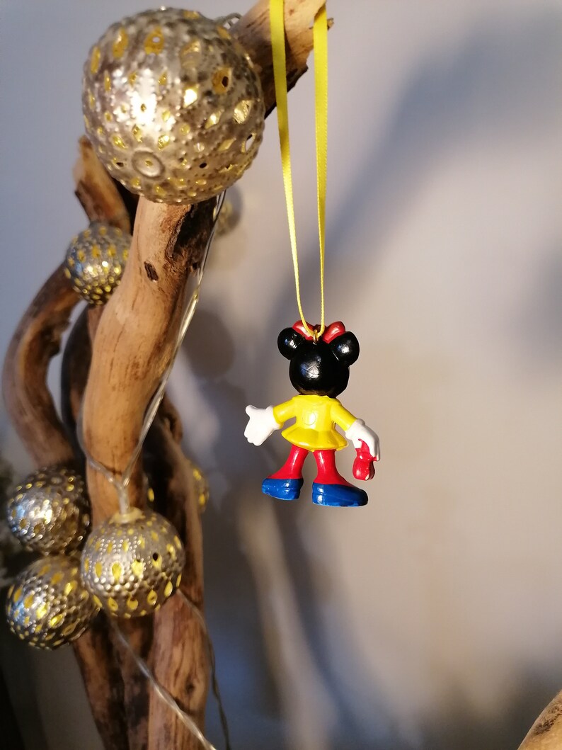 K&ouml;nnte beinhalten: Eine kleine Plastikfigur eines Comic-Charakters, die ein gelbes Kleid, rote Schuhe und eine rote Schleife tr&auml;gt. Die Figur h&auml;ngt an einem gelben Band.