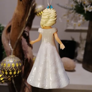 Peut inclure: Une figurine en plastique blanche repr&eacute;sentant une femme aux cheveux blonds portant une robe blanche &agrave; motifs floraux. La figurine est suspendue &agrave; une ficelle blanche.