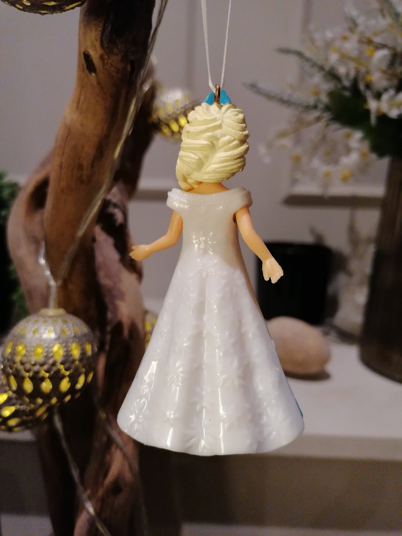 Peut inclure: Une figurine en plastique blanche repr&eacute;sentant une femme aux cheveux blonds portant une robe blanche &agrave; motifs floraux. La figurine est suspendue &agrave; une ficelle blanche.