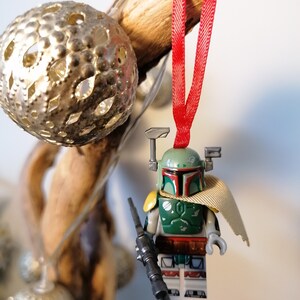 May include: A green and brown Lego Star Wars Boba Fett minifigure ornament with a red ribbon and a black blaster rifle. The minifigure is wearing a tan cape and has a green helmet with a red visor.