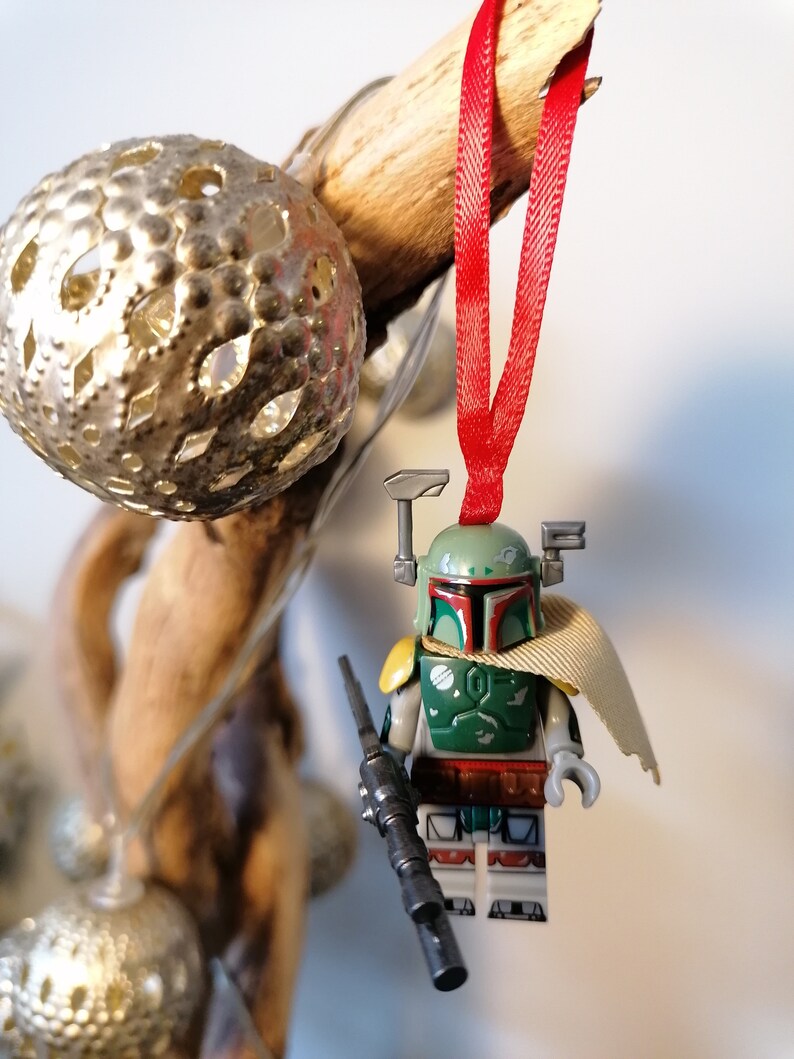 May include: A green and brown Lego Star Wars Boba Fett minifigure ornament with a red ribbon and a black blaster rifle. The minifigure is wearing a tan cape and has a green helmet with a red visor.
