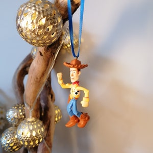 May include: A blue ribbon hangs from a piece of driftwood with a plastic toy figurine of Woody from Toy Story. Woody is wearing his signature cowboy hat, vest, and jeans. He is holding his arm up in the air as if he is waving.