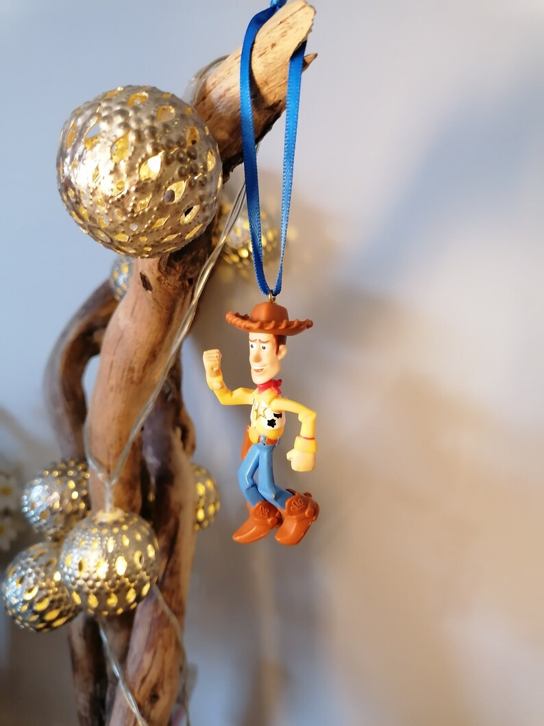May include: A blue ribbon hangs from a piece of driftwood with a plastic toy figurine of Woody from Toy Story. Woody is wearing his signature cowboy hat, vest, and jeans. He is holding his arm up in the air as if he is waving.