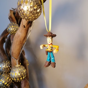 May include: A Woody action figure Christmas ornament with a yellow ribbon. The ornament is hanging in front of a decorative arrangement of silver, spherical lights and driftwood. The Woody figure is wearing a brown cowboy hat, yellow shirt, blue pants, and brown boots.