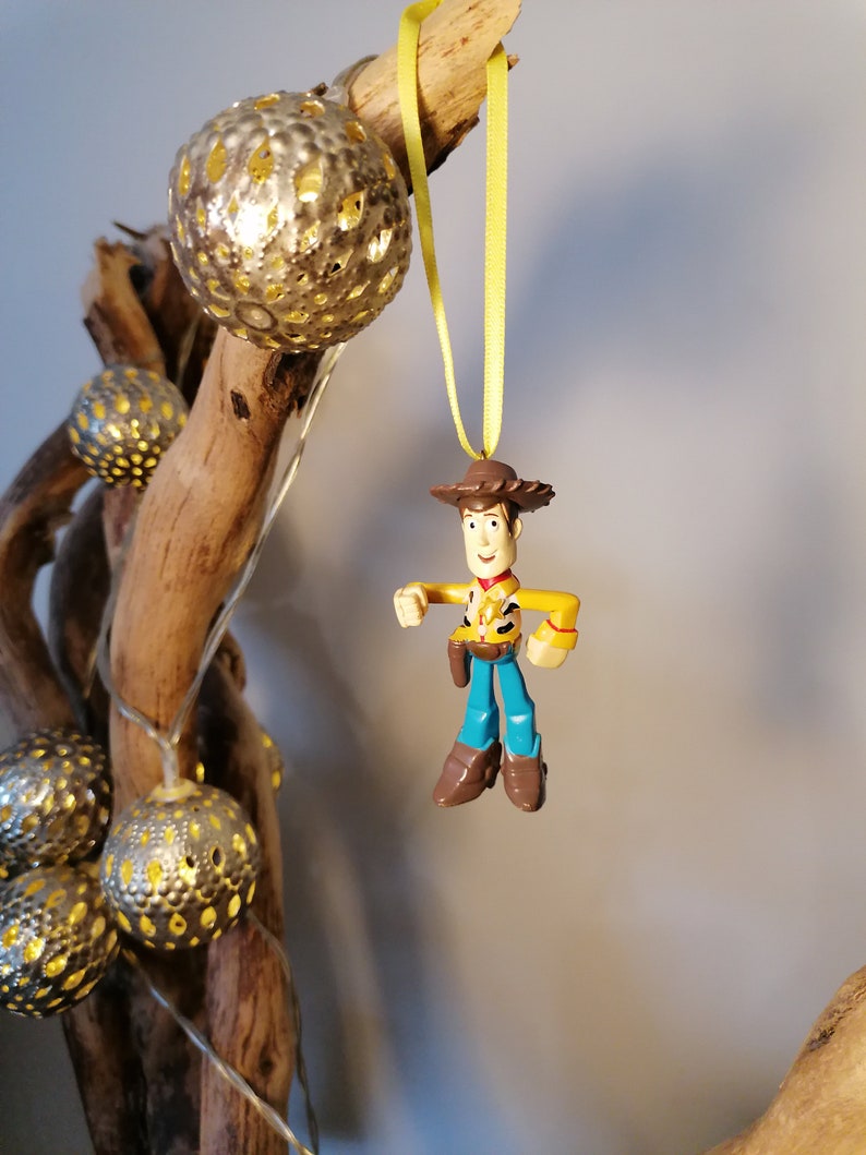 May include: A Woody action figure Christmas ornament with a yellow ribbon. The ornament is hanging in front of a decorative arrangement of silver, spherical lights and driftwood. The Woody figure is wearing a brown cowboy hat, yellow shirt, blue pants, and brown boots.