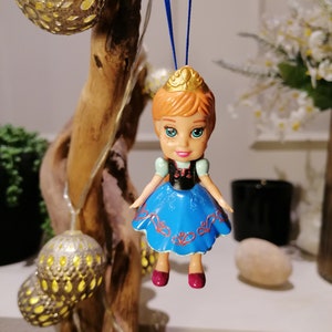 K&ouml;nnte beinhalten: Ein blauer und wei&szlig;er Plastik-Puppen-Anh&auml;nger einer Prinzessin mit einer goldenen Krone, roten Schuhen und einem blauen Kleid mit wei&szlig;en und roten Details. Die Puppe h&auml;ngt an einer blauen Schnur.