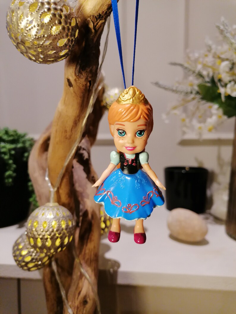 K&ouml;nnte beinhalten: Ein blauer und wei&szlig;er Plastik-Puppen-Anh&auml;nger einer Prinzessin mit einer goldenen Krone, roten Schuhen und einem blauen Kleid mit wei&szlig;en und roten Details. Die Puppe h&auml;ngt an einer blauen Schnur.