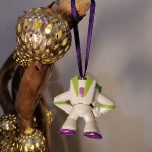 May include: A white plastic toy astronaut ornament with green and purple details hanging from a purple ribbon. The ornament is hanging from a branch with gold and silver ornaments.