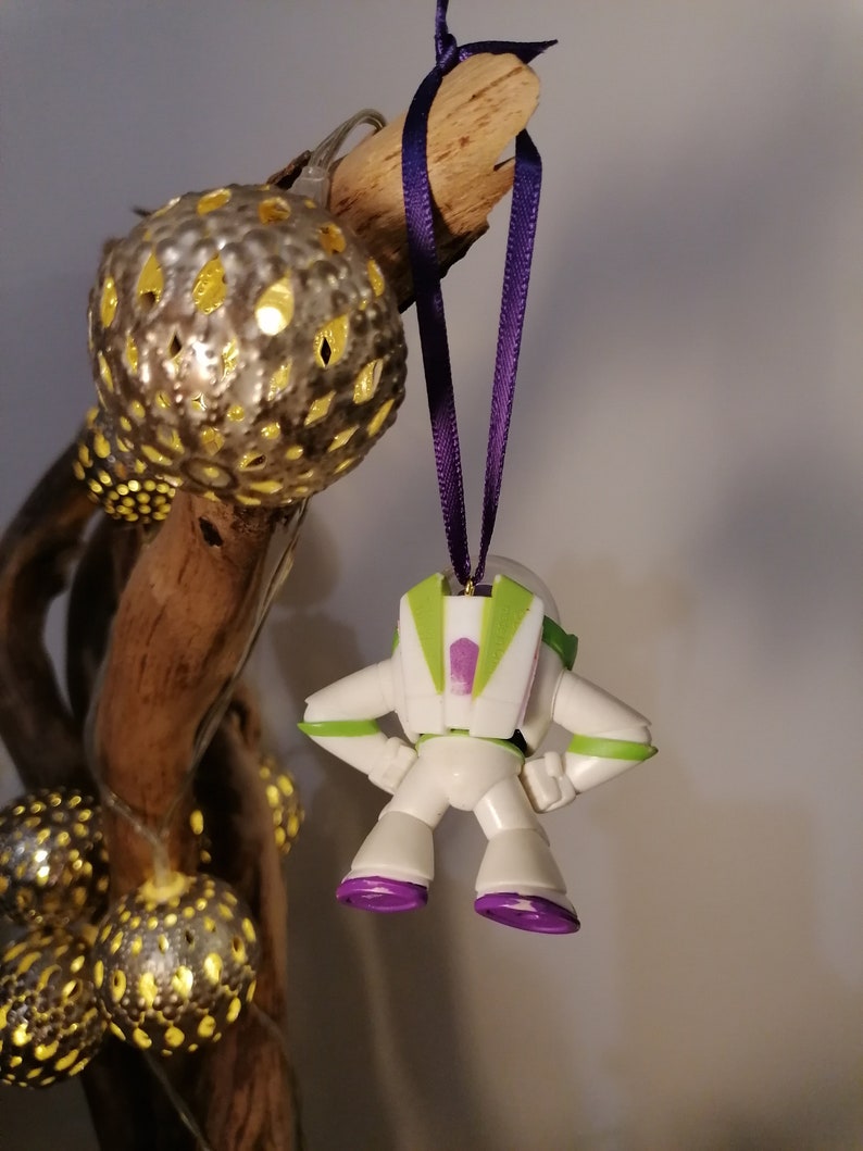 May include: A white plastic toy astronaut ornament with green and purple details hanging from a purple ribbon. The ornament is hanging from a branch with gold and silver ornaments.