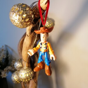 May include: A red and brown cowboy figurine ornament with a red ribbon hanging from a branch. The figurine is wearing a brown hat, a yellow vest, blue jeans, and brown boots.