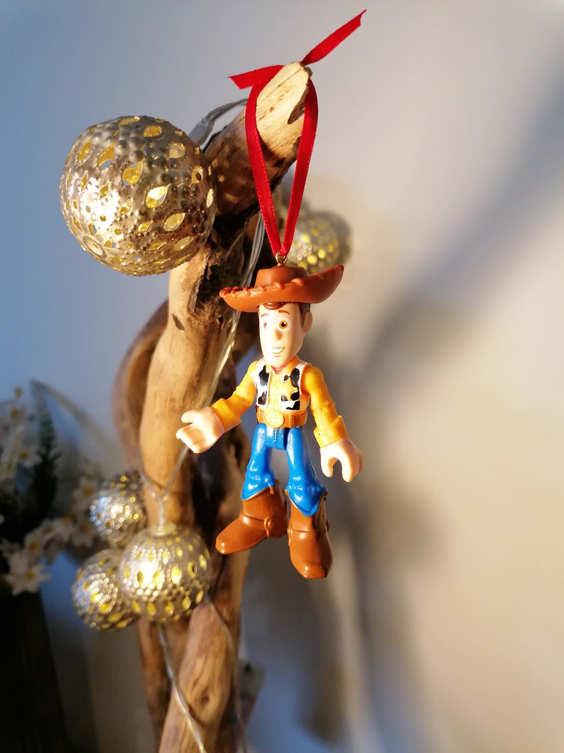 May include: A red and brown cowboy figurine ornament with a red ribbon hanging from a branch. The figurine is wearing a brown hat, a yellow vest, blue jeans, and brown boots.