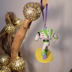 K&ouml;nnte beinhalten: Ein wei&szlig;er und gr&uuml;ner Buzz Lightyear Weihnachtsbaumschmuck, der an einem Ast mit einem violetten Band h&auml;ngt. Der Schmuck ist in Form von Buzz Lightyear aus dem Film Toy Story, und er steht auf einem gelben Surfbrett.