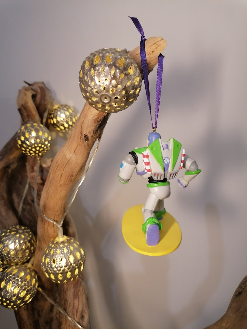 K&ouml;nnte beinhalten: Ein wei&szlig;er und gr&uuml;ner Buzz Lightyear Weihnachtsbaumschmuck, der an einem Ast mit einem violetten Band h&auml;ngt. Der Schmuck ist in Form von Buzz Lightyear aus dem Film Toy Story, und er steht auf einem gelben Surfbrett.