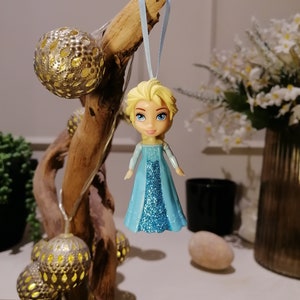 May include: A blue and silver glitter bauble featuring a cartoon character with blonde hair and a blue dress. The bauble is hanging from a blue ribbon.