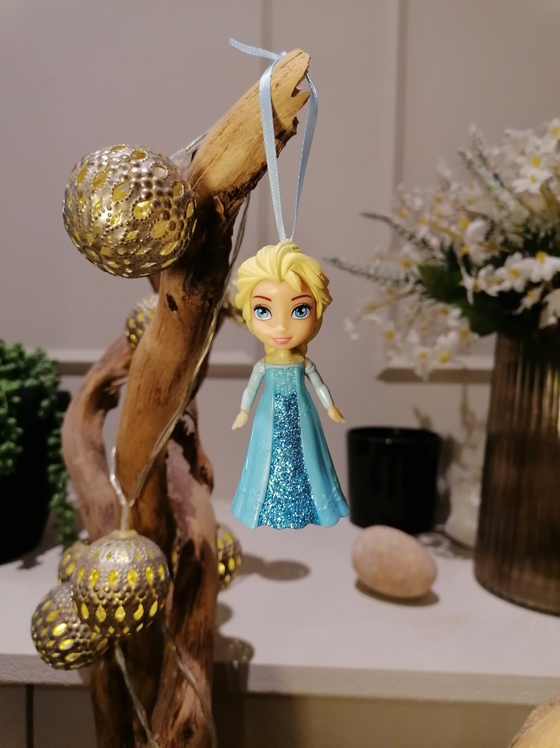 May include: A blue and silver glitter bauble featuring a cartoon character with blonde hair and a blue dress. The bauble is hanging from a blue ribbon.
