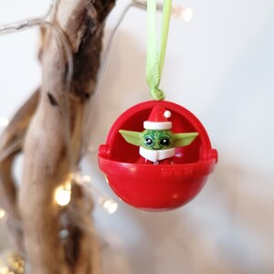 Pu&ograve; includere: Un ornamento natalizio rosso con una piccola figura verde e bianca di un personaggio che indossa un cappello da Babbo Natale all'interno. L'ornamento &egrave; appeso a un nastro verde.