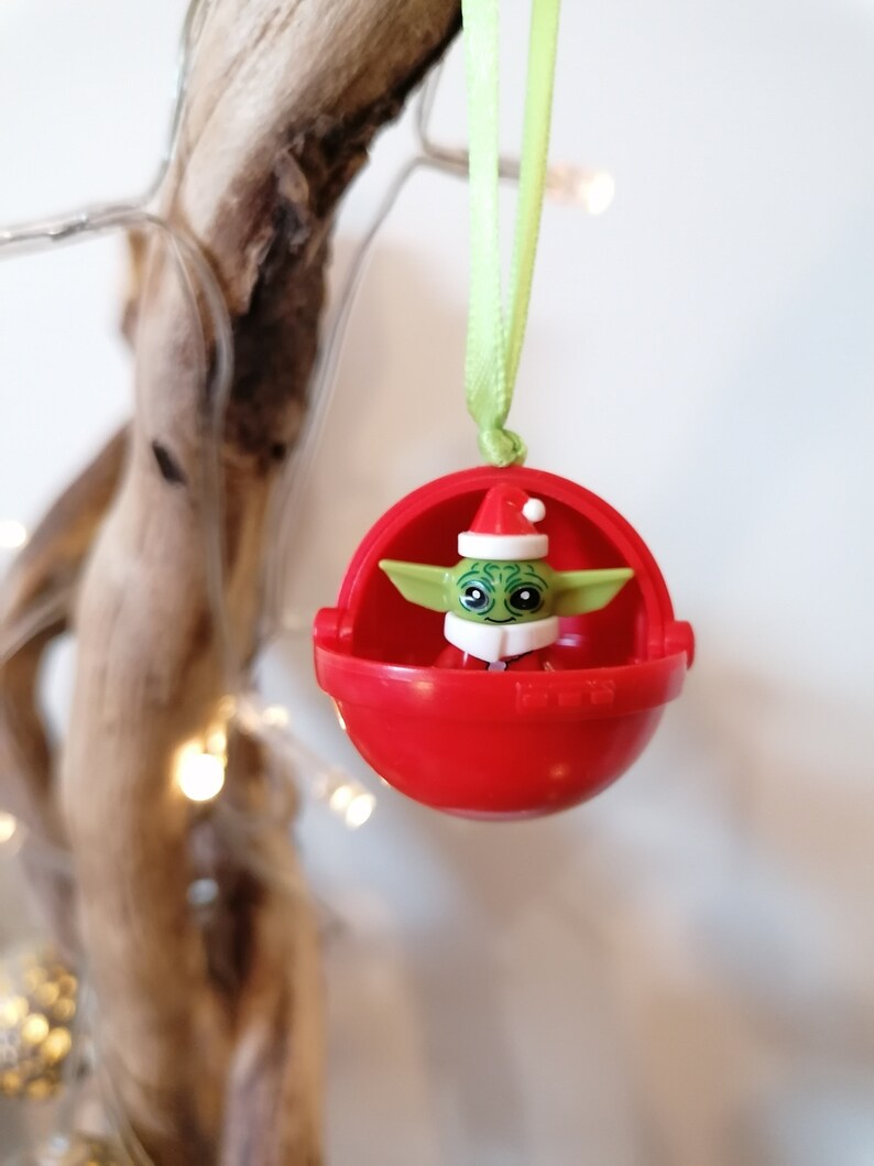 Pu&ograve; includere: Un ornamento natalizio rosso con una piccola figura verde e bianca di un personaggio che indossa un cappello da Babbo Natale all'interno. L'ornamento &egrave; appeso a un nastro verde.