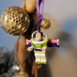 May include: A purple and white Christmas bauble featuring a Lego figurine of Buzz Lightyear from the Toy Story film franchise. The bauble is hanging from a purple ribbon.