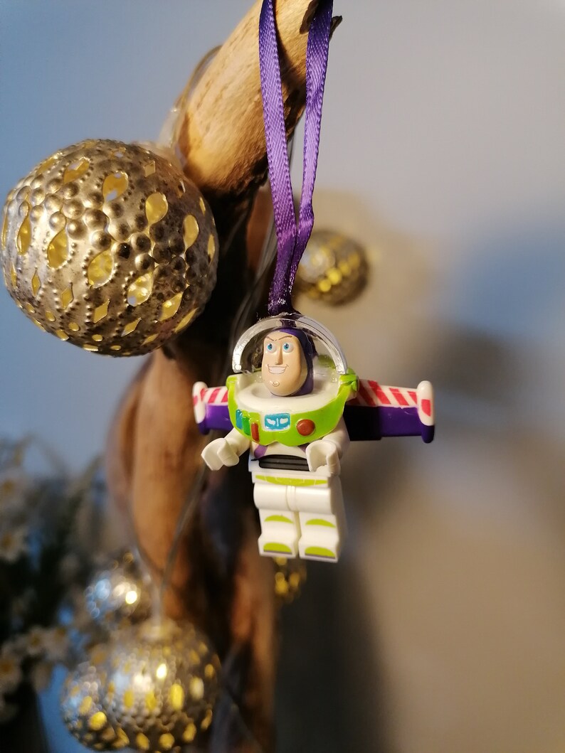 May include: A purple and white Christmas bauble featuring a Lego figurine of Buzz Lightyear from the Toy Story film franchise. The bauble is hanging from a purple ribbon.