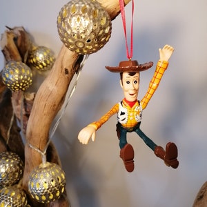 May include: A plastic ornament of Woody from Toy Story, wearing his signature cowboy hat and vest, with a red string attached. He is hanging from a branch with gold-colored, decorative balls.