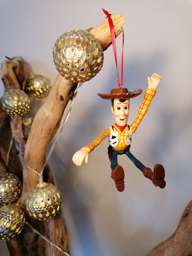 May include: A plastic ornament of Woody from Toy Story, wearing his signature cowboy hat and vest, with a red string attached. He is hanging from a branch with gold-colored, decorative balls.