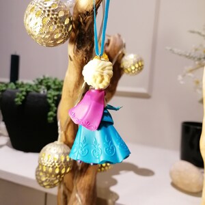 K&ouml;nnte beinhalten: Ein blauer und rosa Plastik-Anh&auml;nger mit einer Prinzessin mit einer wei&szlig;en Krone und blonden Haaren. Der Anh&auml;nger h&auml;ngt an einem blauen Band an einem Holzast.
