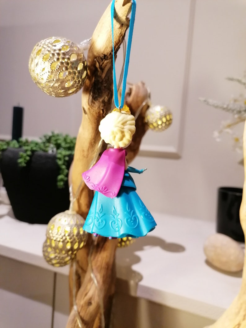 K&ouml;nnte beinhalten: Ein blauer und rosa Plastik-Anh&auml;nger mit einer Prinzessin mit einer wei&szlig;en Krone und blonden Haaren. Der Anh&auml;nger h&auml;ngt an einem blauen Band an einem Holzast.
