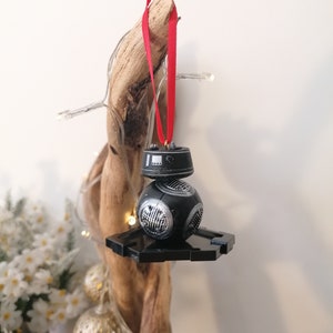 K&ouml;nnte beinhalten: Schwarzer und silberner Star Wars BB-8 Weihnachtsbaumschmuck mit einem roten Band, das an einem St&uuml;ck Treibholz h&auml;ngt.