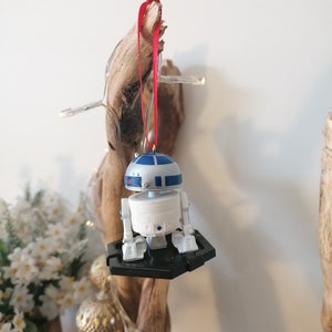 May include: A white and blue R2-D2 Star Wars character ornament hanging from a red ribbon on a wooden branch. The ornament is attached to a black plastic base.
