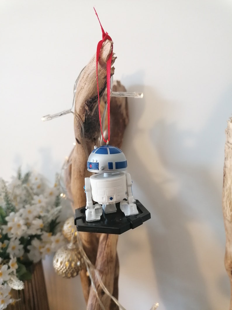 May include: A white and blue R2-D2 Star Wars character ornament hanging from a red ribbon on a wooden branch. The ornament is attached to a black plastic base.