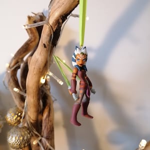 May include: A green and white Star Wars character ornament with green lightsabers. The ornament is hanging from a green ribbon on a branch.