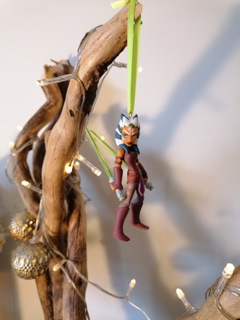 May include: A green and white Star Wars character ornament with green lightsabers. The ornament is hanging from a green ribbon on a branch.