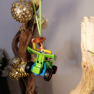May include: A green and blue toy car with a figure of Woody from Toy Story driving it. The car is hanging from a branch with a green ribbon.