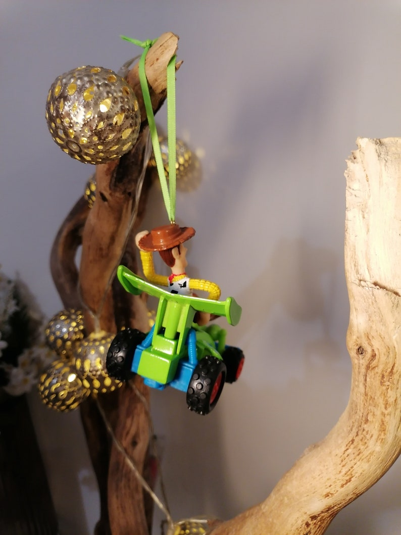 May include: A green and blue toy car with a figure of Woody from Toy Story driving it. The car is hanging from a branch with a green ribbon.