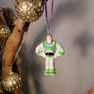 Puede incluir: Un adorno de plástico blanco y verde de Buzz Lightyear de la franquicia Toy Story. El adorno está colgado de una cinta morada.