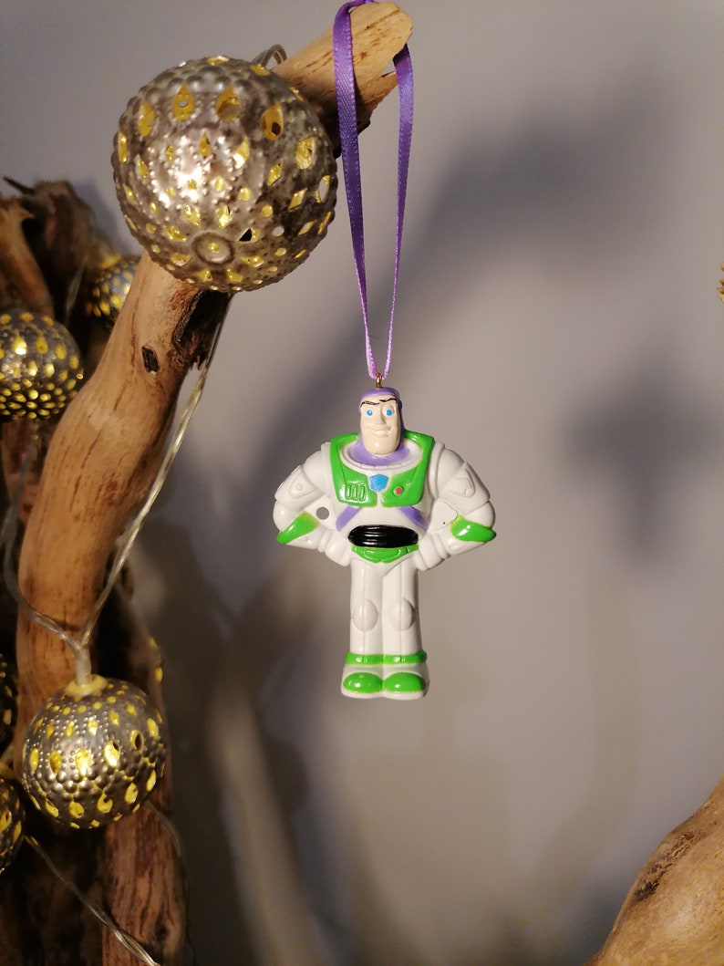 Puede incluir: Un adorno de pl&aacute;stico blanco y verde de Buzz Lightyear de la franquicia Toy Story. El adorno est&aacute; colgado de una cinta morada.