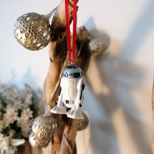 K&ouml;nnte beinhalten: Ein wei&szlig;er und blauer R2-D2 Star Wars Charakter-Anh&auml;nger, der an einem roten Band an einem Treibholzast mit goldenen und silbernen Ornamenten h&auml;ngt.