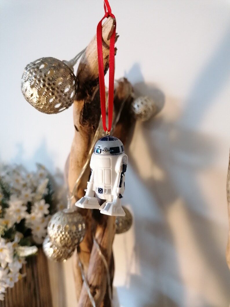 K&ouml;nnte beinhalten: Ein wei&szlig;er und blauer R2-D2 Star Wars Charakter-Anh&auml;nger, der an einem roten Band an einem Treibholzast mit goldenen und silbernen Ornamenten h&auml;ngt.