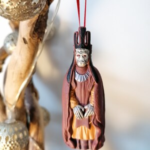 May include: A red and brown Christmas ornament depicting a character from Star Wars. The character is wearing a crown and a long robe. The ornament is hanging from a red ribbon.
