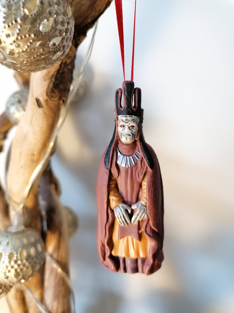 May include: A red and brown Christmas ornament depicting a character from Star Wars. The character is wearing a crown and a long robe. The ornament is hanging from a red ribbon.