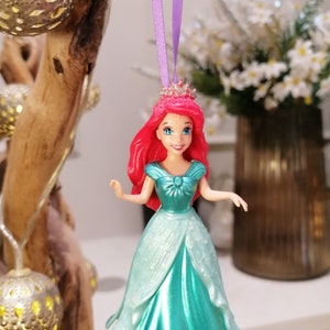 May include: A red-haired princess doll wearing a turquoise gown with glitter accents. She is wearing a crown and has a purple ribbon attached to her head. The doll is hanging from a tree branch.
